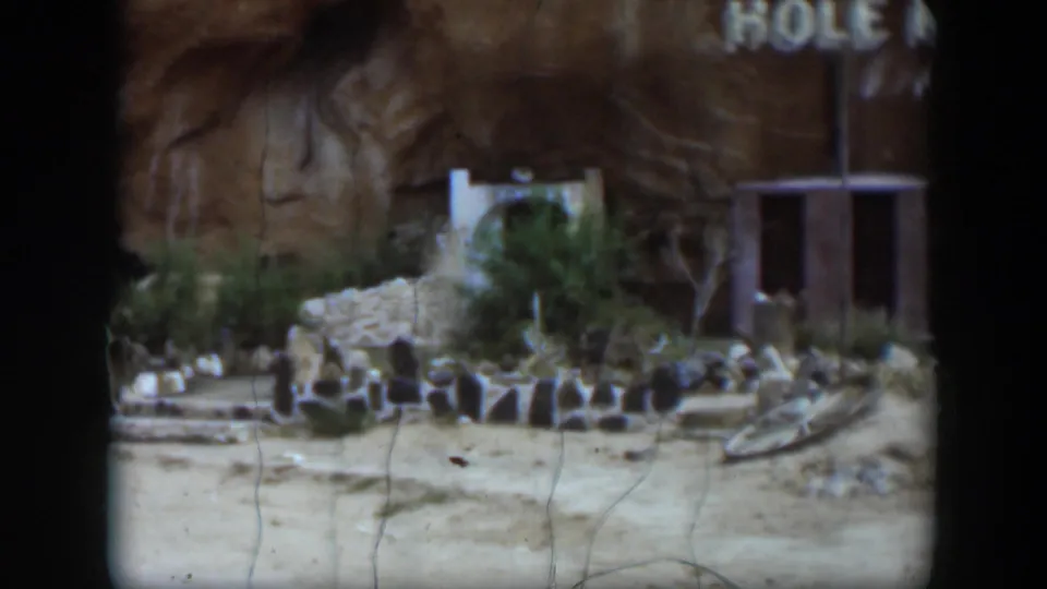 The final frame holds on the Utah ruin area, showing low stone remains, sandy ground, dry vegetation, a partial sign, and a small structure at right.