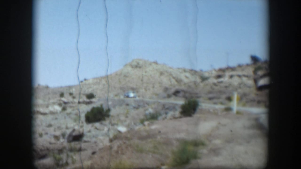 1951 Utah desert road scene with rocky slopes, sparse shrubs, distant cars, and visible 8mm film scratches.