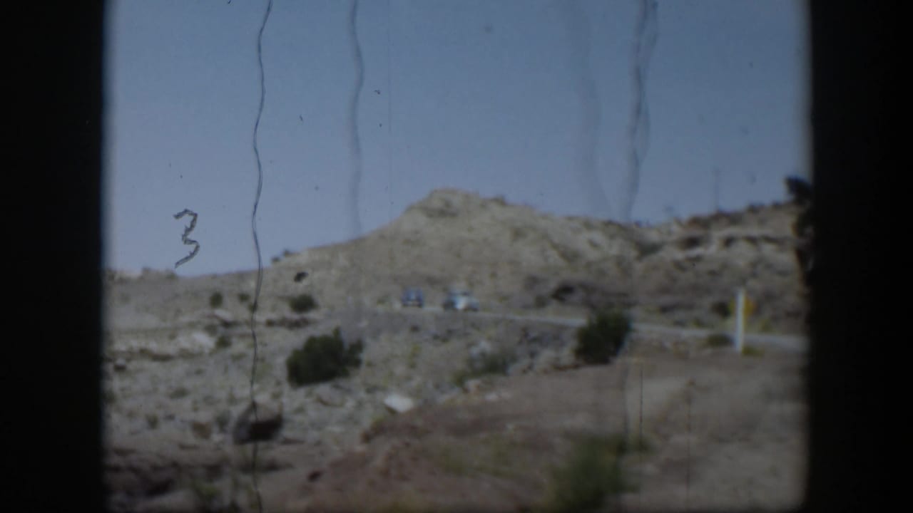 1951 Utah road-travel frame showing arid hills, sparse brush, distant vehicles, and dark vehicle-window borders.
