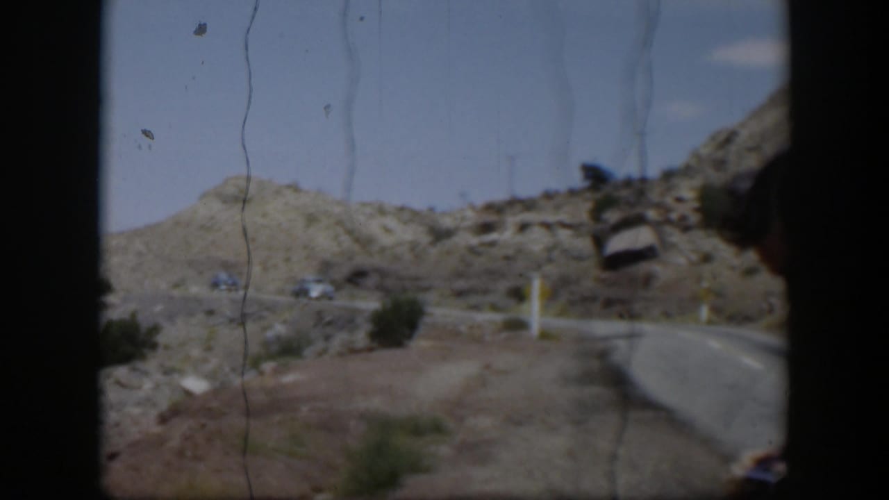 1951 Utah home-movie frame with a curving paved road, rocky desert terrain, shrubs, and distant cars under blue sky.