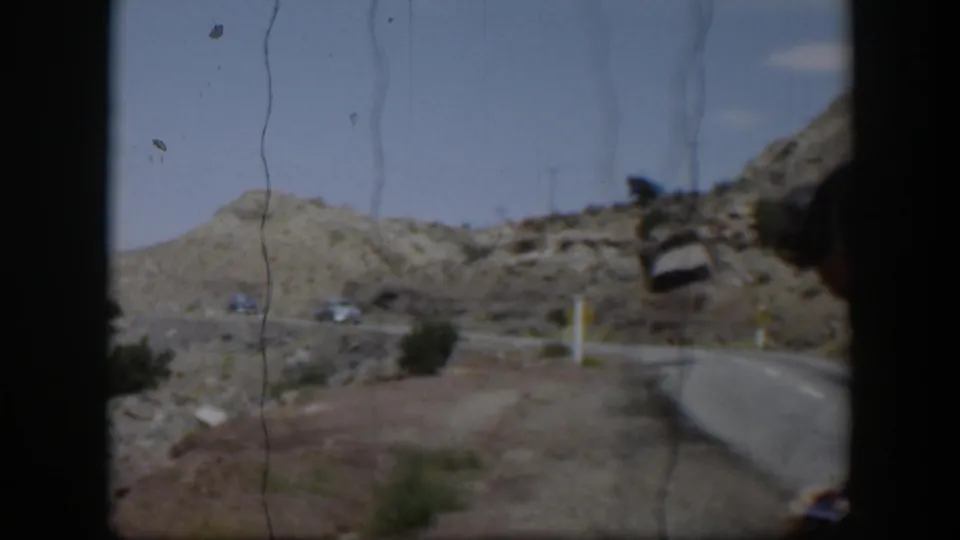 1951 Utah home-movie frame with a curving paved road, rocky desert terrain, shrubs, and distant cars under blue sky.
