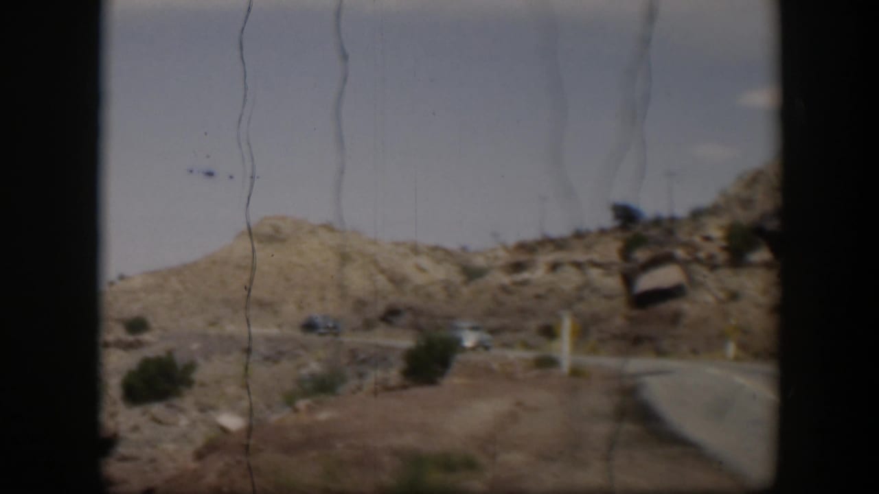 A moving-vehicle view of a curving Utah desert road in 1951, with distant cars, hills, shrubs, and visible film wear.