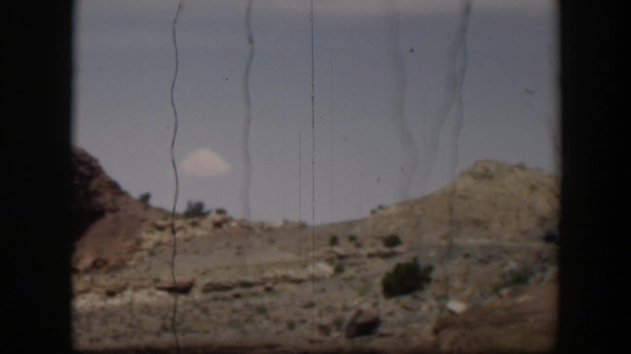 Utah desert hills and roadside scrub appear from a moving 8mm home-movie viewpoint, with a small cloud and visible film scratches.