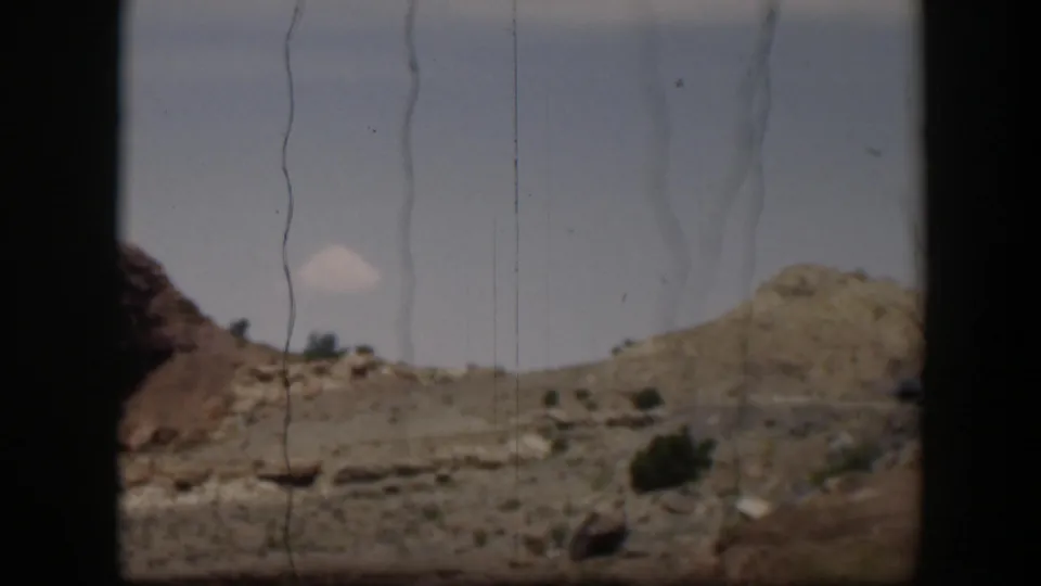 Utah desert hills and roadside scrub appear from a moving 8mm home-movie viewpoint, with a small cloud and visible film scratches.