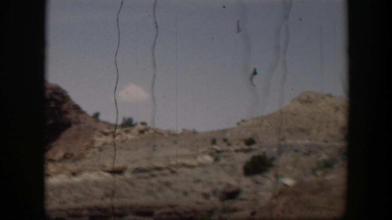 A 1951 Utah desert roadside view with rocky hills, sparse shrubs, pale sky, and prominent 8mm scratches and frame borders.