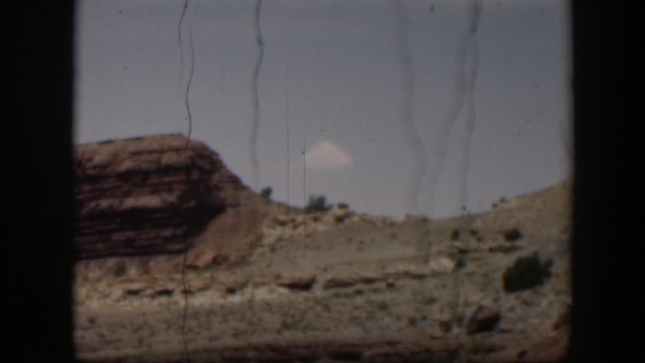 Layered red rock and pale desert hillside under a bright sky, seen through dark film-frame edges.