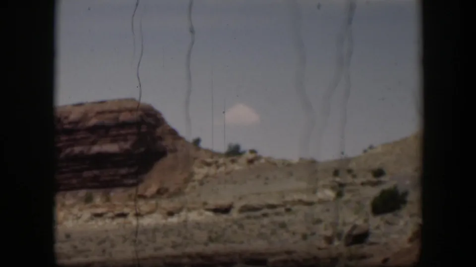 1951 Utah home-movie frame showing layered desert rock, scrubby hillside, scattered stones, and a bright sky through a dark-edged view.