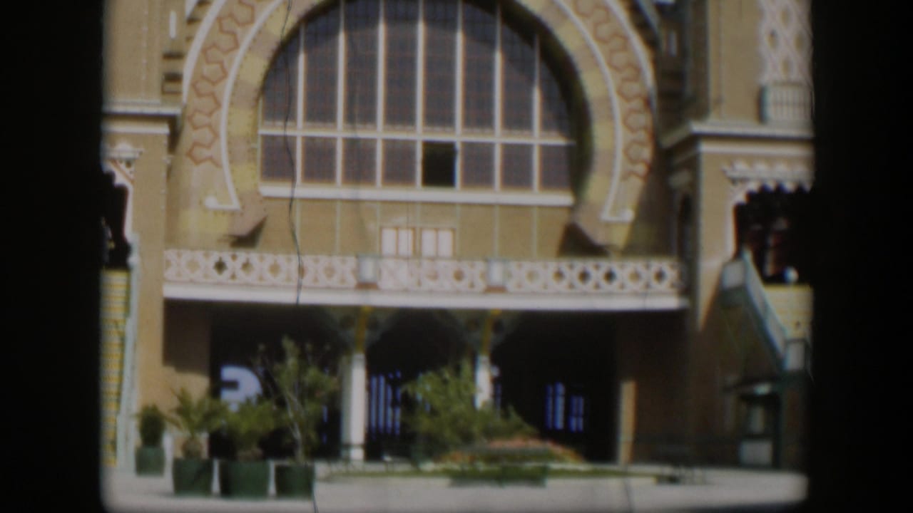 1951 Utah 8mm frame with an ornate tan building front, arched window, white lattice detail, potted plants, and shaded entrances.