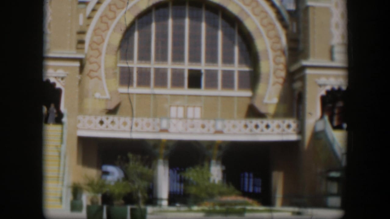 1951 Utah 8mm frame showing the tan arched-window facade, exterior stairway, white balcony trim, planted base, and a partial vehicle.