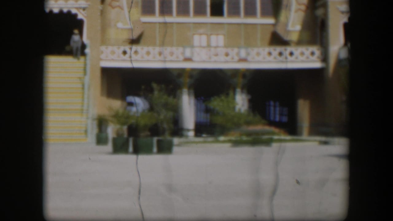 1951 Utah home-movie frame cropped across the yellow stairs, white trim, lower arches, potted plants, and a distant person near the doorway.