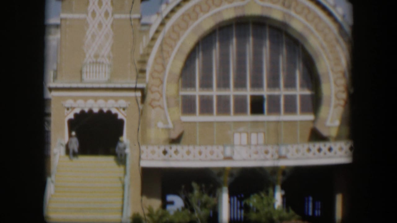 1951 Utah 8mm frame showing a large arched window, ornate yellow facade, white trim, broad stairs, plants, and two distant figures.