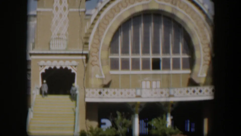 1951 Utah 8mm frame showing a large arched window, ornate yellow facade, white trim, broad stairs, plants, and two distant figures.