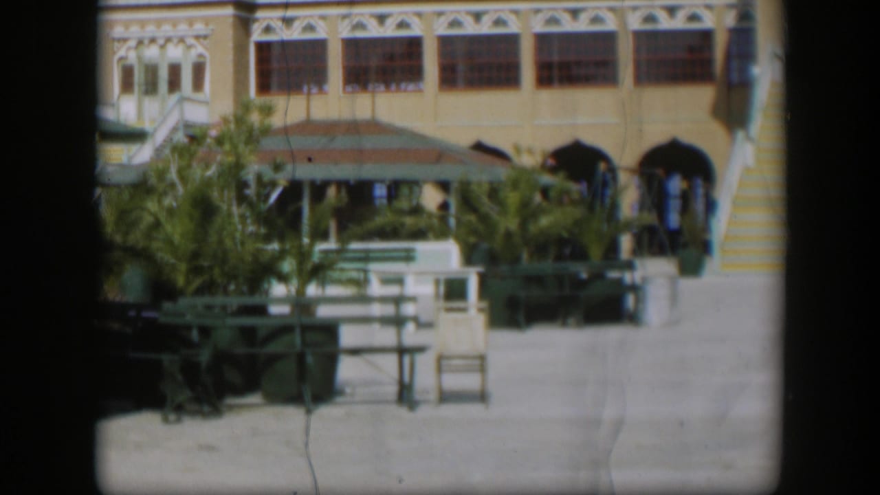1951 home-movie frame of a Utah patio with green benches, palms, arched doorways, decorative trim, and small people near the entrance.