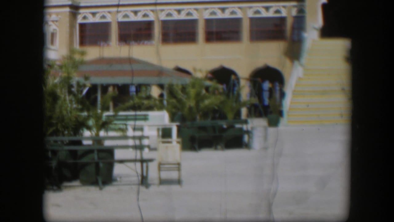 Sunlit 1951 Utah exterior with yellow-striped stairs, arched doorways, green benches, umbrellas, plants, and distant people by the entrance.