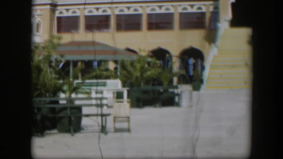 Sunlit 1951 Utah exterior with yellow-striped stairs, arched doorways, green benches, umbrellas, plants, and distant people by the entrance.