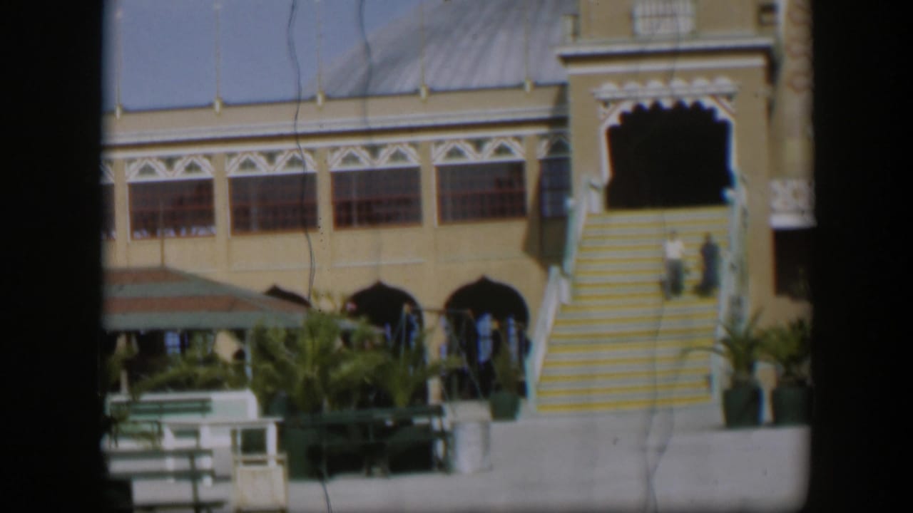 1951 Utah footage frame with a resort facade, yellow-and-white steps, courtyard benches, potted plants and two distant visitors on the stairs.