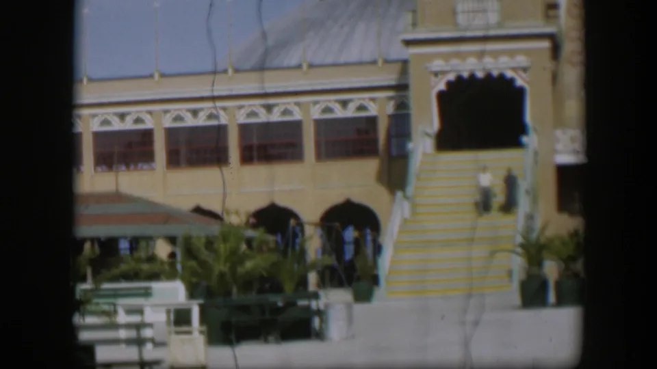 1951 Utah footage frame with a resort facade, yellow-and-white steps, courtyard benches, potted plants and two distant visitors on the stairs.