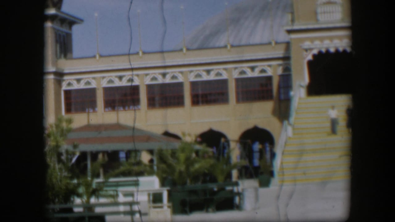 1951 Utah home-movie frame showing the resort courtyard from a closer angle, with striped steps, arches, benches and plants in bright sun.