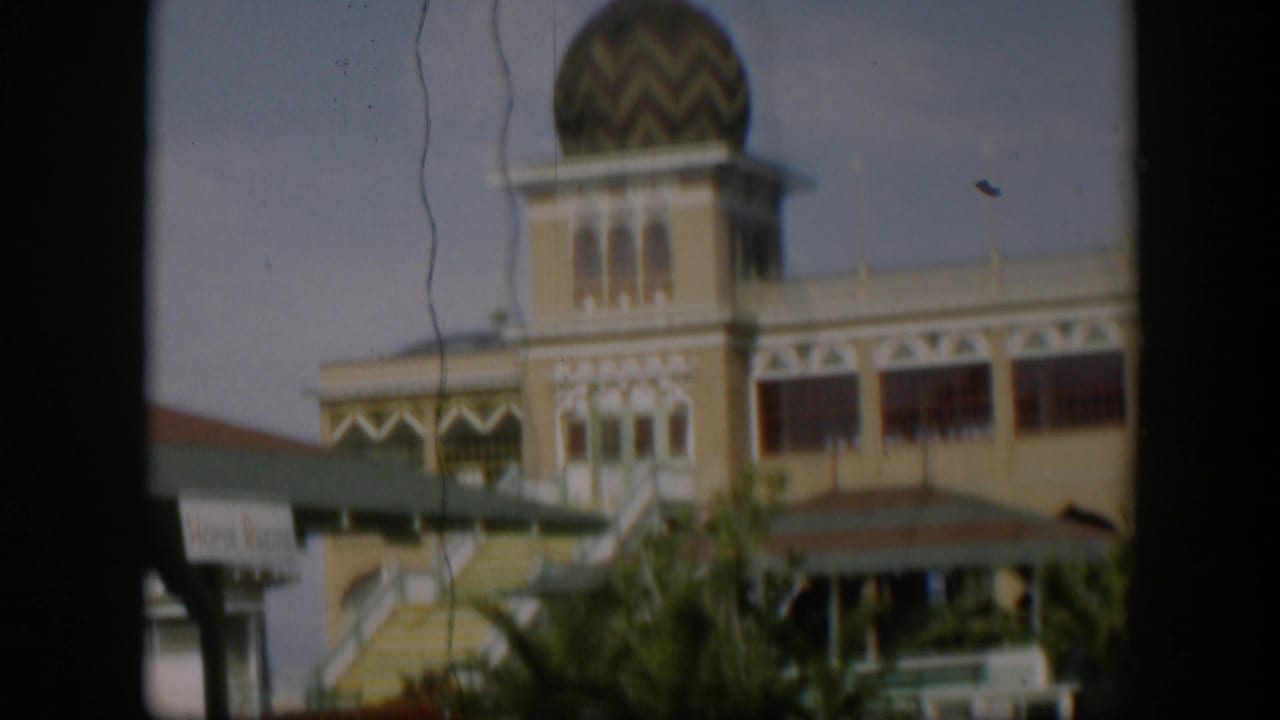 1951 Utah 8mm frame showing a yellow building with a domed tower, exterior stairs, green awning, and plants viewed through a dark frame.