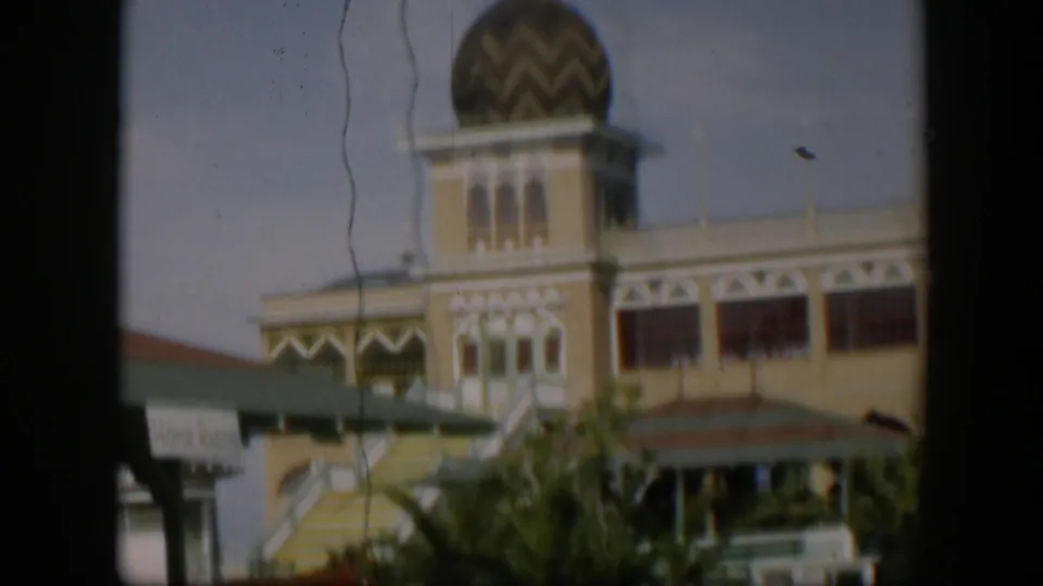 1951 Utah 8mm frame showing a yellow building with a domed tower, exterior stairs, green awning, and plants viewed through a dark frame.
