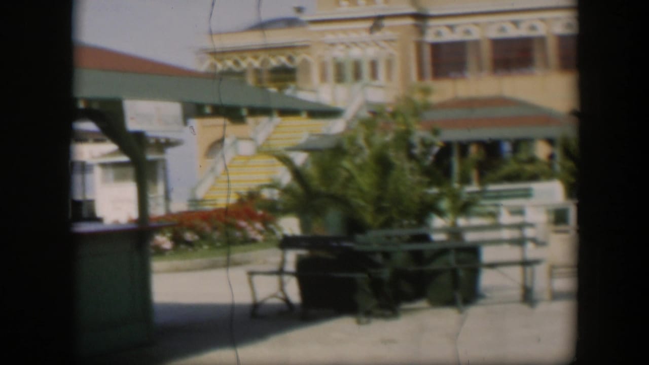 1951 Utah home-movie frame showing the yellow building beyond plants, railings, a green awning, exterior stairs, and benches or tables.