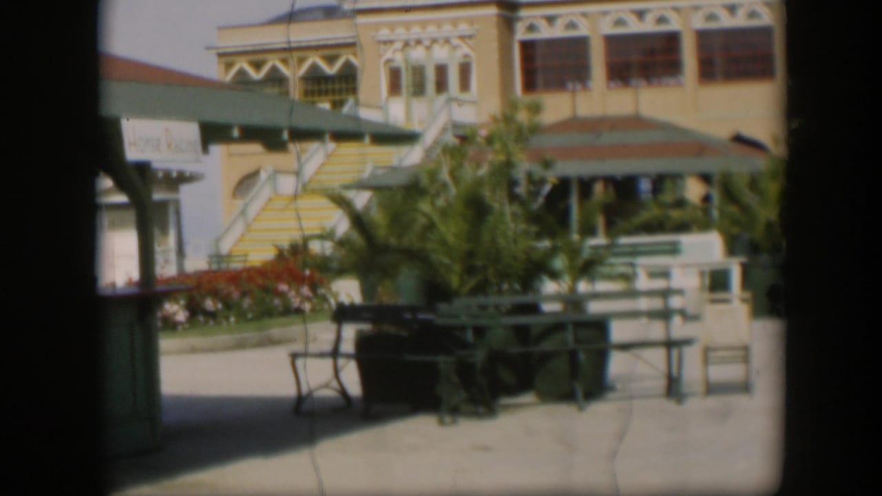 1951 Utah home-movie view of a yellow resort courtyard with striped stairs, green benches, palms, and people near a shaded entry.