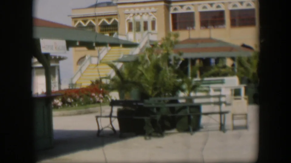 1951 Utah home-movie view of a yellow resort courtyard with striped stairs, green benches, palms, and people near a shaded entry.