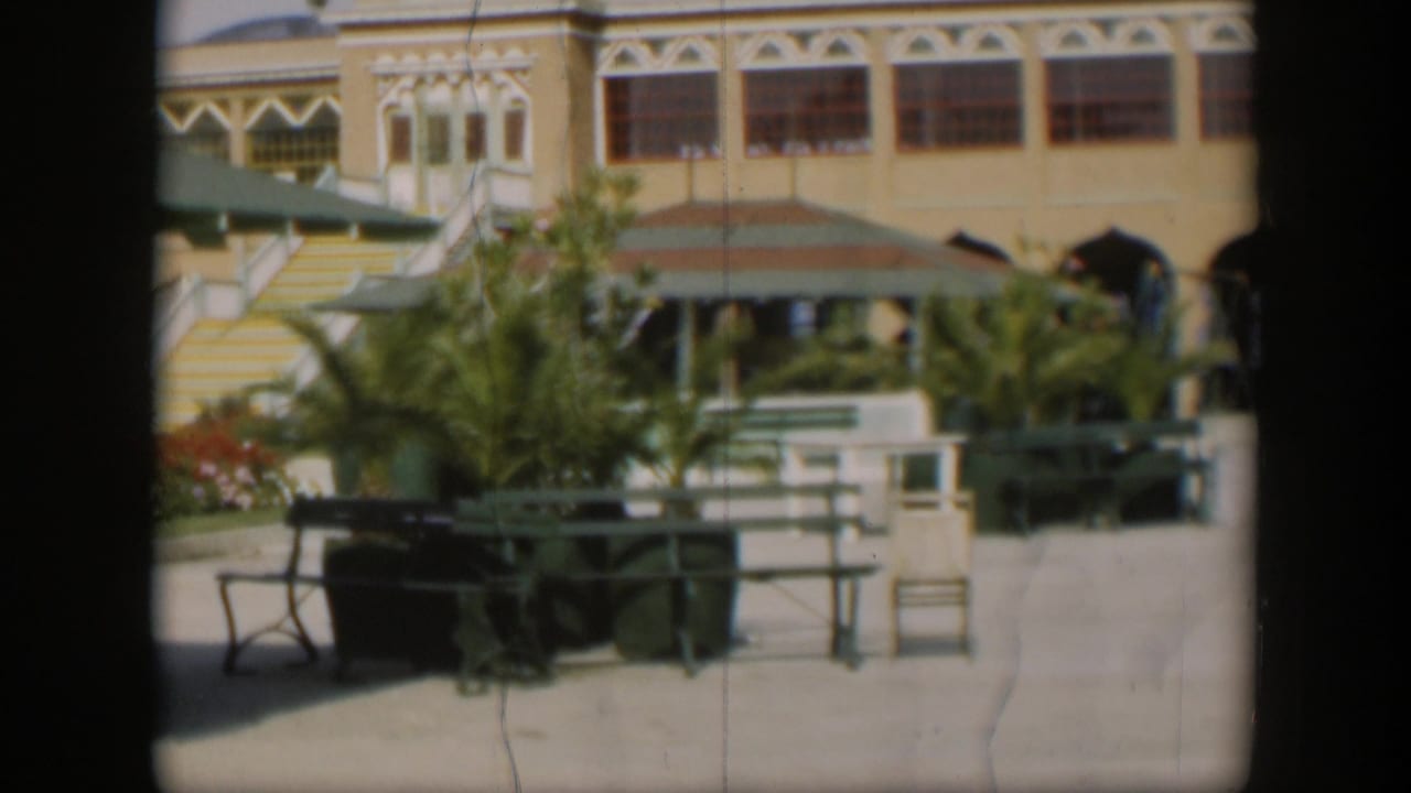 1951 Utah resort courtyard with green benches, palms, yellow facade, striped stairs, and distant visitors near the entrance.
