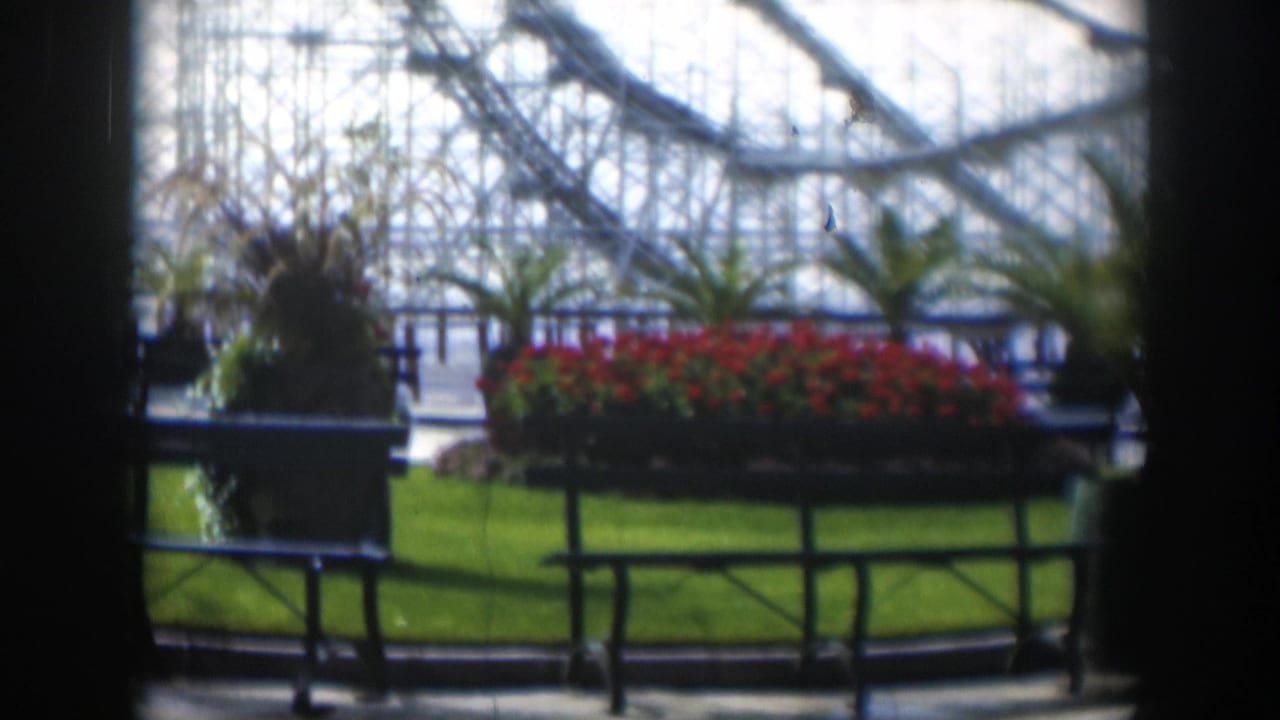 1951 Utah amusement park view through a dark interior frame, showing red planters, green grass, railings, and a roller coaster structure.