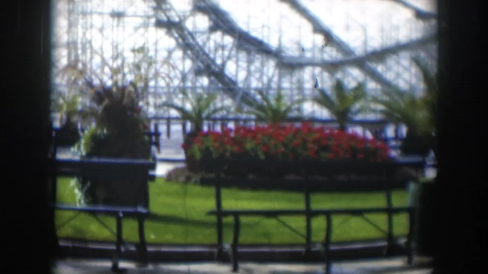 1951 Utah amusement park view through a dark interior frame, showing red planters, green grass, railings, and a roller coaster structure.