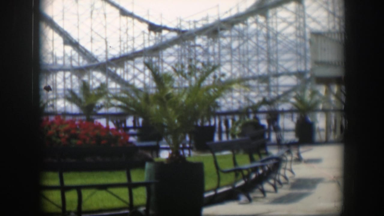 1951 Utah amusement park frame showing a potted palm, red flower bed, empty benches, paved walkway, and coaster structure in the background.