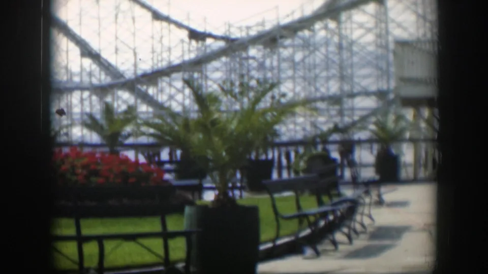 1951 Utah amusement park frame showing a potted palm, red flower bed, empty benches, paved walkway, and coaster structure in the background.