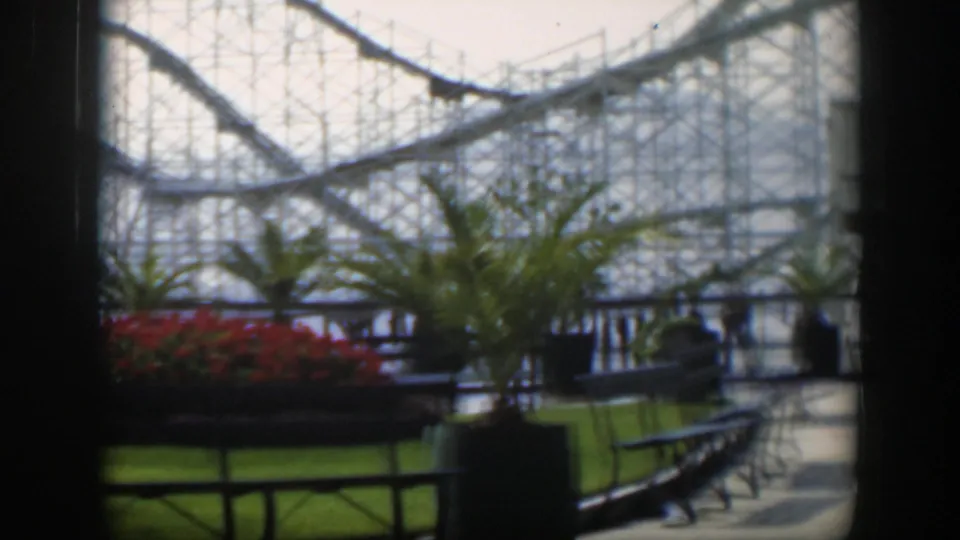 1951 Utah 8mm view of a roller coaster behind palms, red flowers, lawn, railings and benches from a shaded vantage point.