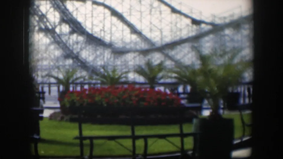 1951 Utah amusement park view with roller coaster supports, palm planters, red flowers, railings and a lawn area in soft 8mm focus.