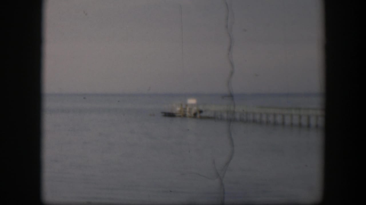 1951 Utah home-movie frame with a low, quiet view of a wooden pier stretching across calm water under pale cloud cover.