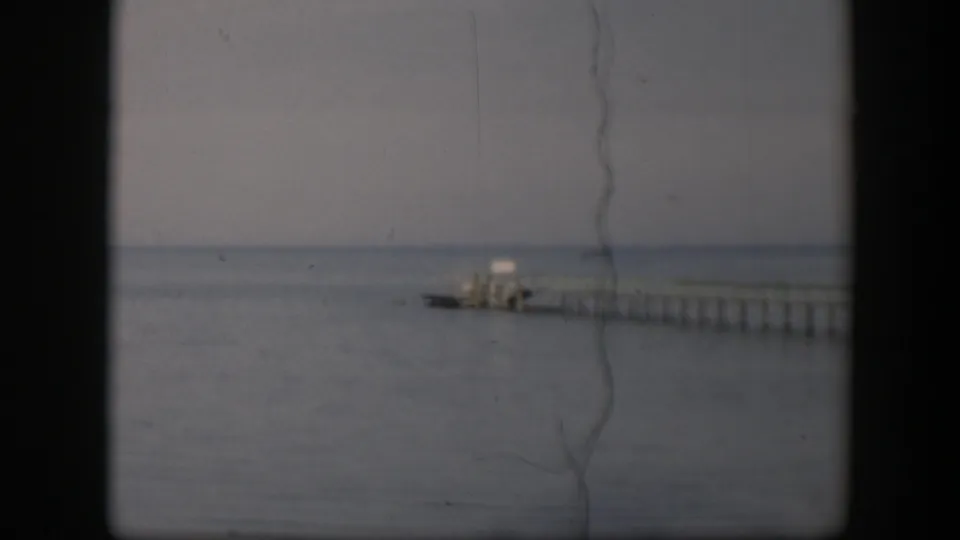 1951 Utah home-movie frame with a low, quiet view of a wooden pier stretching across calm water under pale cloud cover.