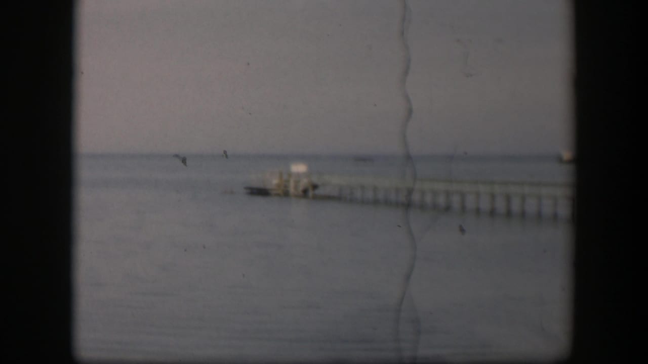 Muted 1951 Utah 8mm frame showing the pier receding over blue-grey water, with visible grain and vertical scratch marks.