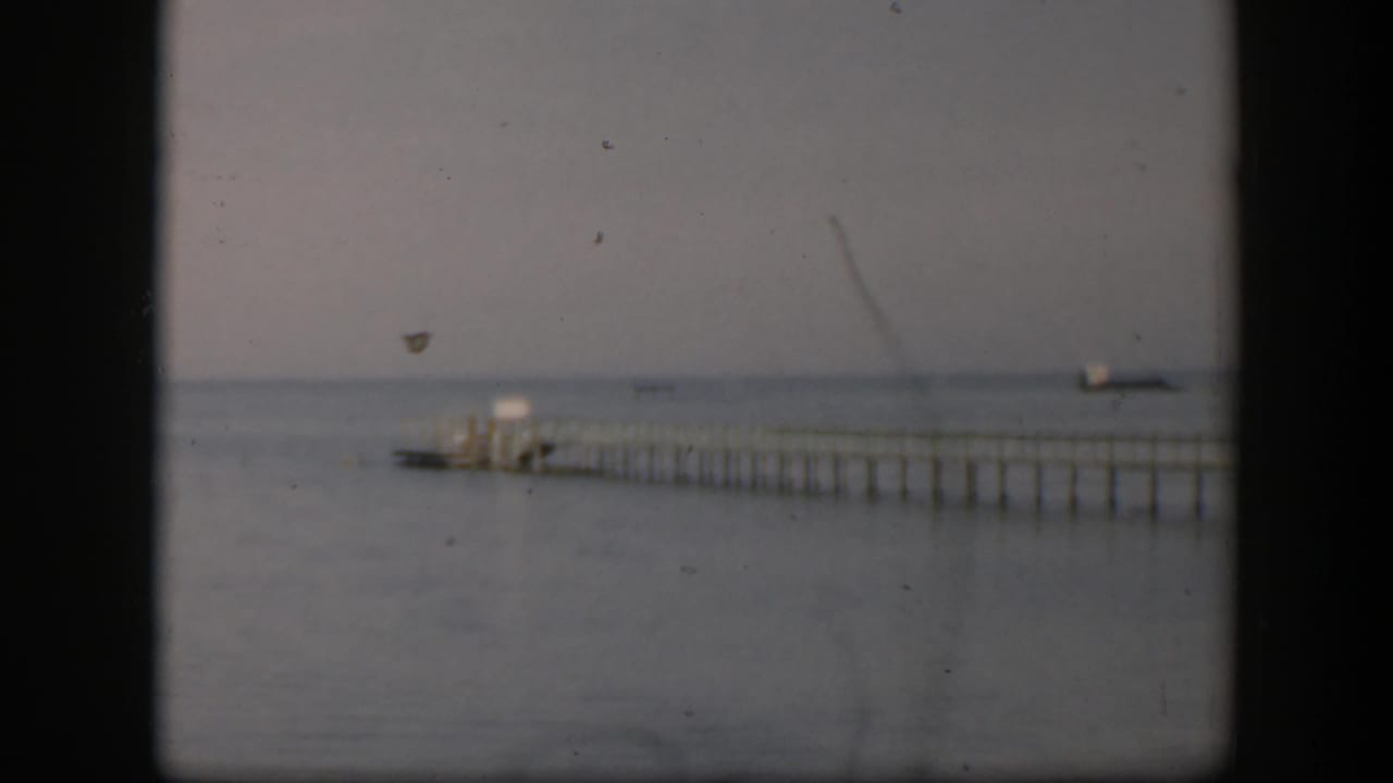 1951 Utah home-movie frame with a long pier, small boat near the dock end, calm water, and soft overcast light.