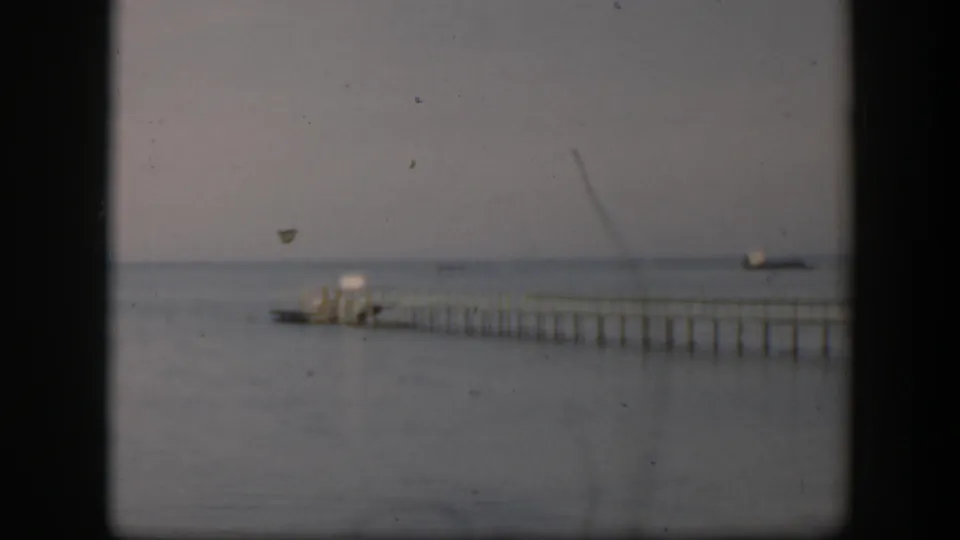1951 Utah home-movie frame with a long pier, small boat near the dock end, calm water, and soft overcast light.