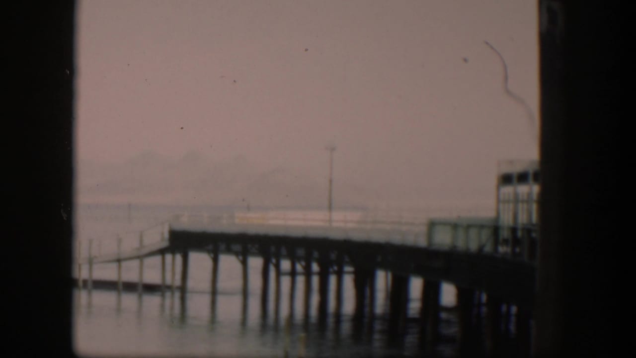 1951 Utah home-movie frame with the pier end or dock section at right, calm water below, and a hazy shoreline or landform in the background.