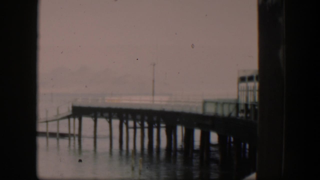 Wooden dock extends over calm water beneath a hazy sky with dark film borders.
