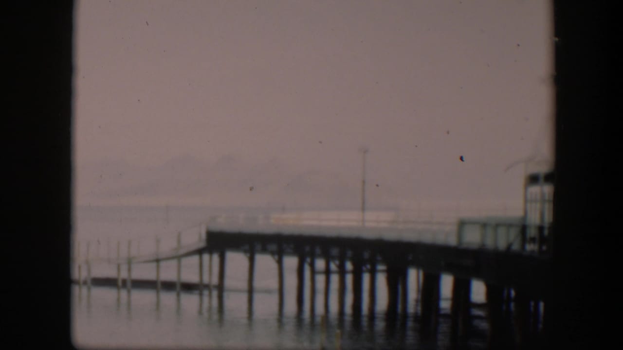 1951 Utah home-movie frame with a dark-edged view of a quiet dock, pale lake water, and mountains softened by mist.