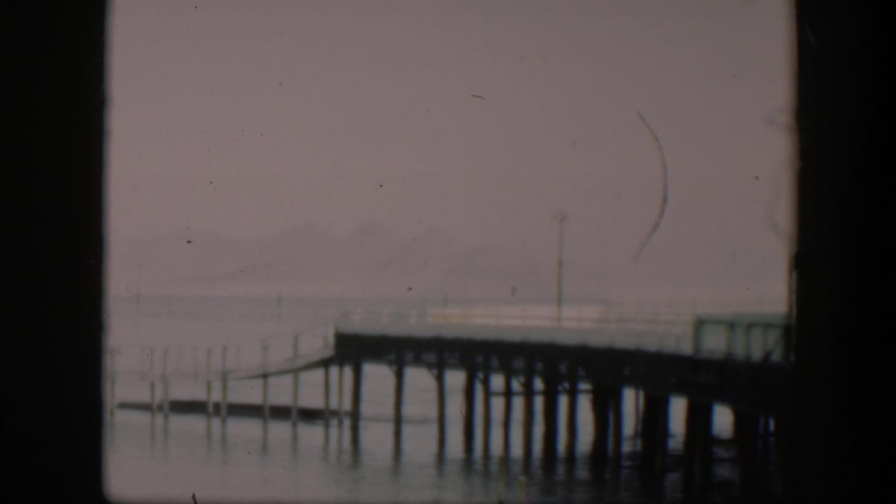 1951 Utah 8mm frame showing the dock from a slightly lower composition, with pilings reflected in calm water and a hazy mountain backdrop.