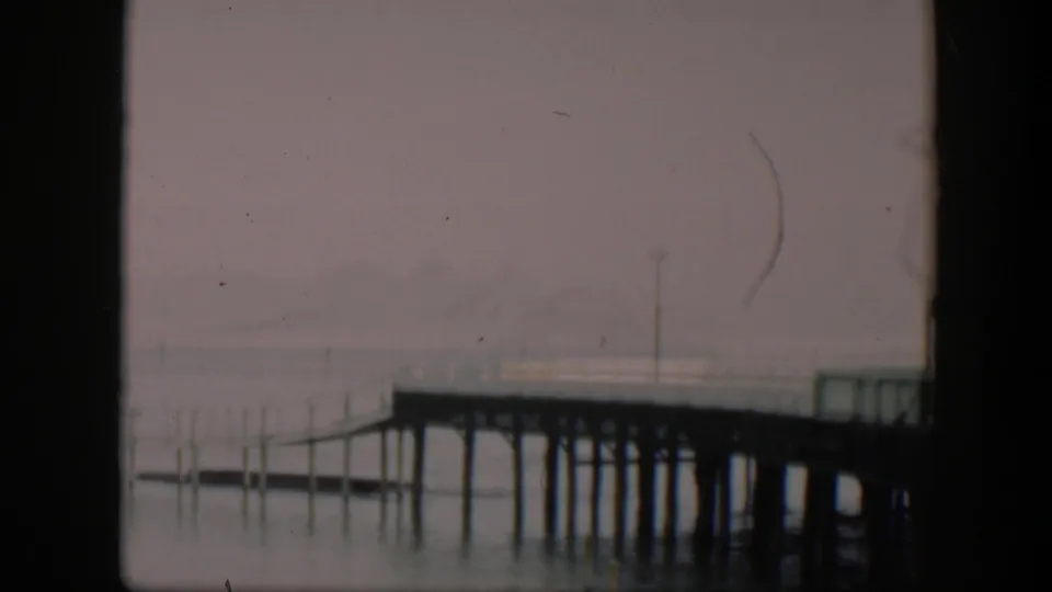 1951 Utah 8mm frame showing the dock from a slightly lower composition, with pilings reflected in calm water and a hazy mountain backdrop.