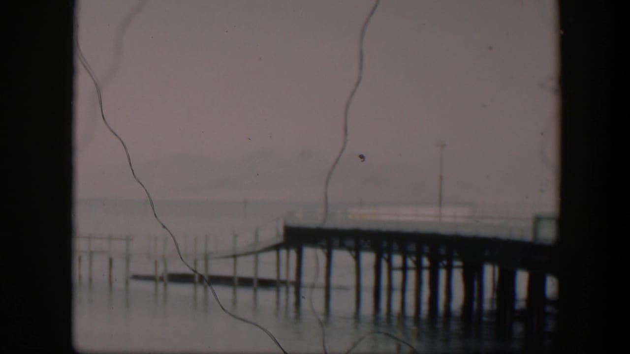 1951 Utah 8mm frame with a wooden dock crossing misty lake water, dark frame edges, and prominent film scratches over the image.