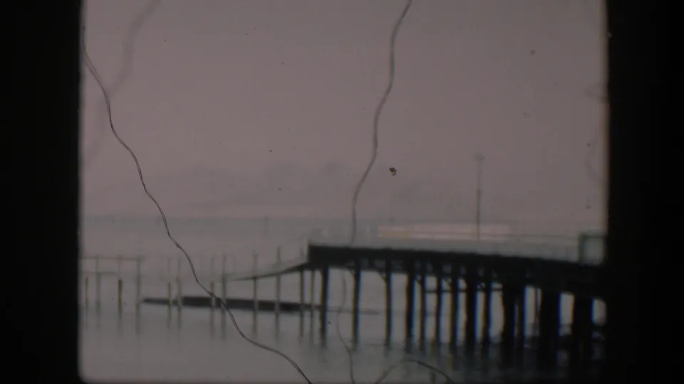 1951 Utah 8mm frame with a wooden dock crossing misty lake water, dark frame edges, and prominent film scratches over the image.