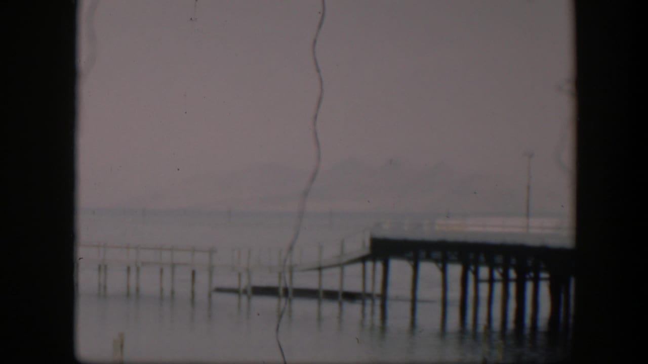 A steady 1951 Utah waterfront view with a wooden pier, scattered posts in the water, muted sky, and visible film wear.
