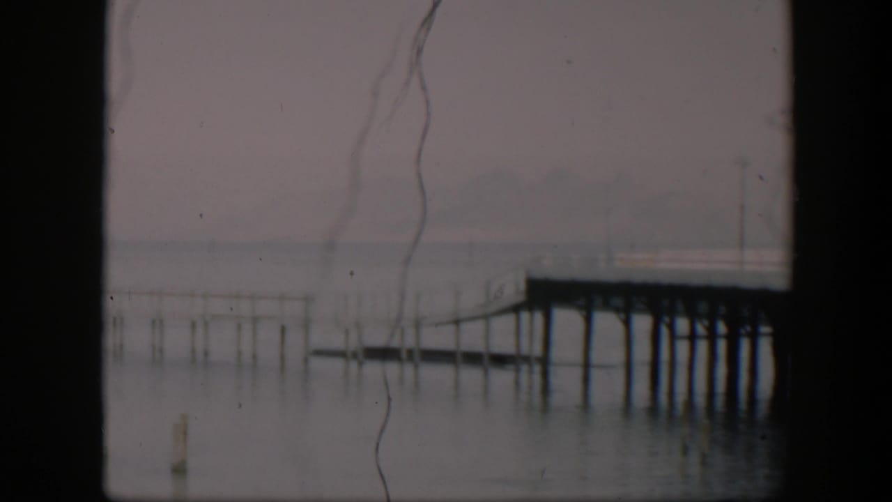 Faded 8mm frame from 1951 Utah metadata showing a quiet pier scene with calm water, thin posts, and soft distant landforms.