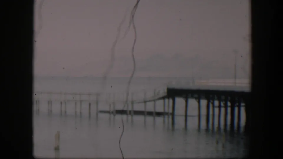 Faded 8mm frame from 1951 Utah metadata showing a quiet pier scene with calm water, thin posts, and soft distant landforms.