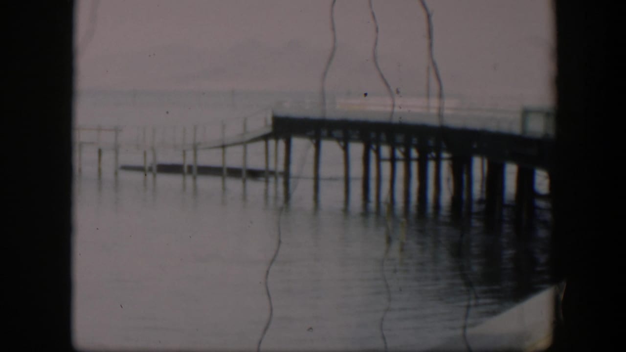A 1951 Utah waterfront frame shows pier supports over calm water, a muted horizon, and pronounced vertical scratches from the 8mm source.